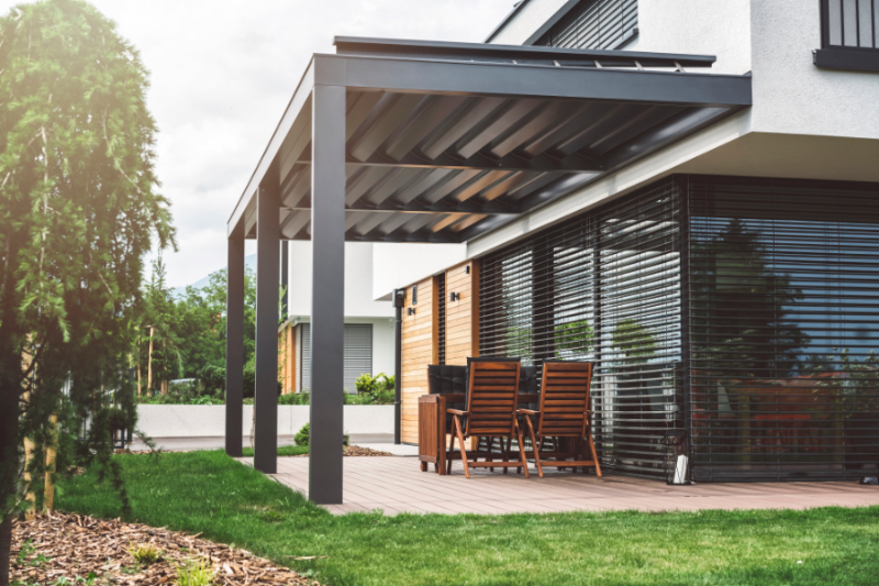 Modern backyard in the suburbs, big windows, wooden chairs and a modern picnic table, white facade of the modern building exterior, beauty of the nature and an amazing upper class residential district.